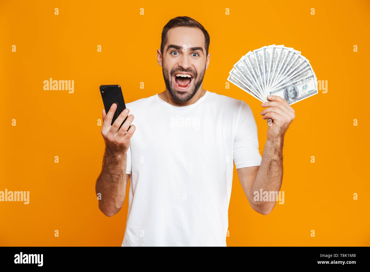 Foto de guapo 30s en ropa casual la celebración de teléfono celular y ventilador de dinero aislado sobre fondo amarillo Foto de stock