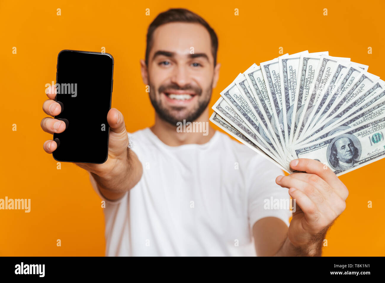 Foto de hombre europeo 30s en ropa casual la celebración de teléfono celular y ventilador de dinero aislado sobre fondo amarillo Foto de stock