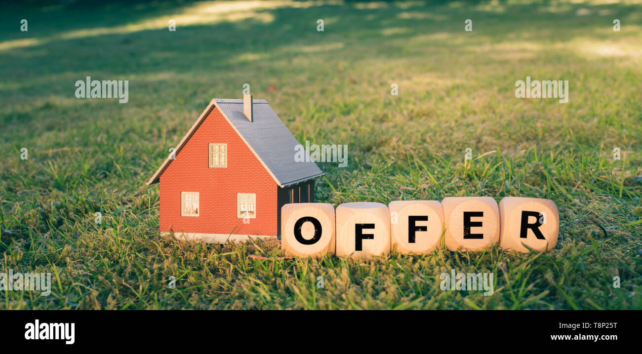 Concepto de hacer una oferta por una casa. Una mano sostiene un modelo casa por encima de una pradera. Dados forman la palabra "oferta". Foto de stock