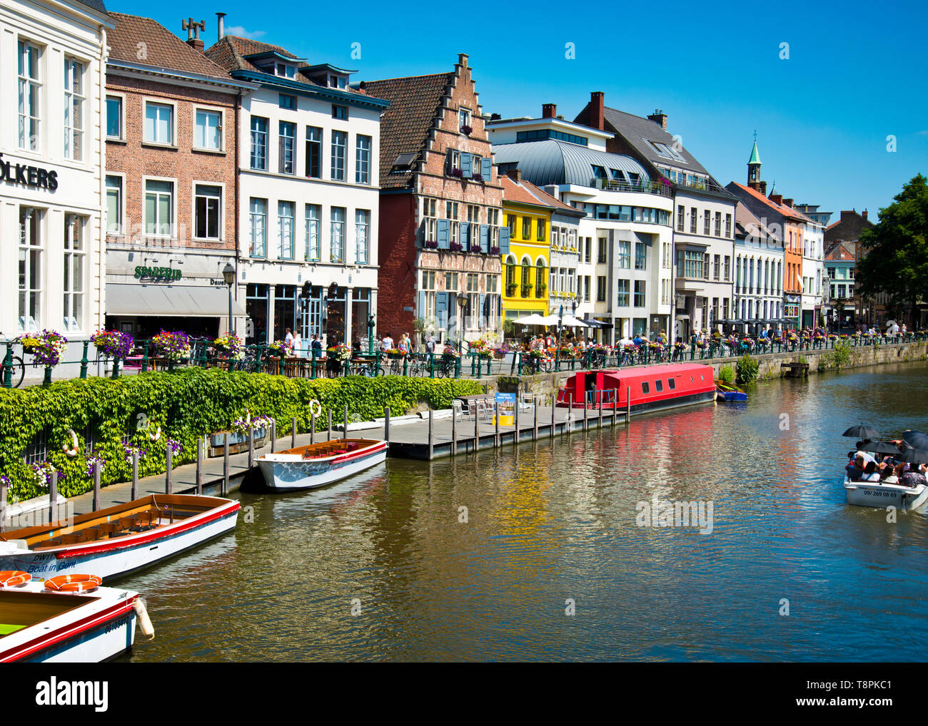 El banco del canal en Gante está lleno de barcos, cafeterías y bares llenos de gente socializando juntos, en un cálido día de verano viendo pasar excursiones en barco. Foto de stock