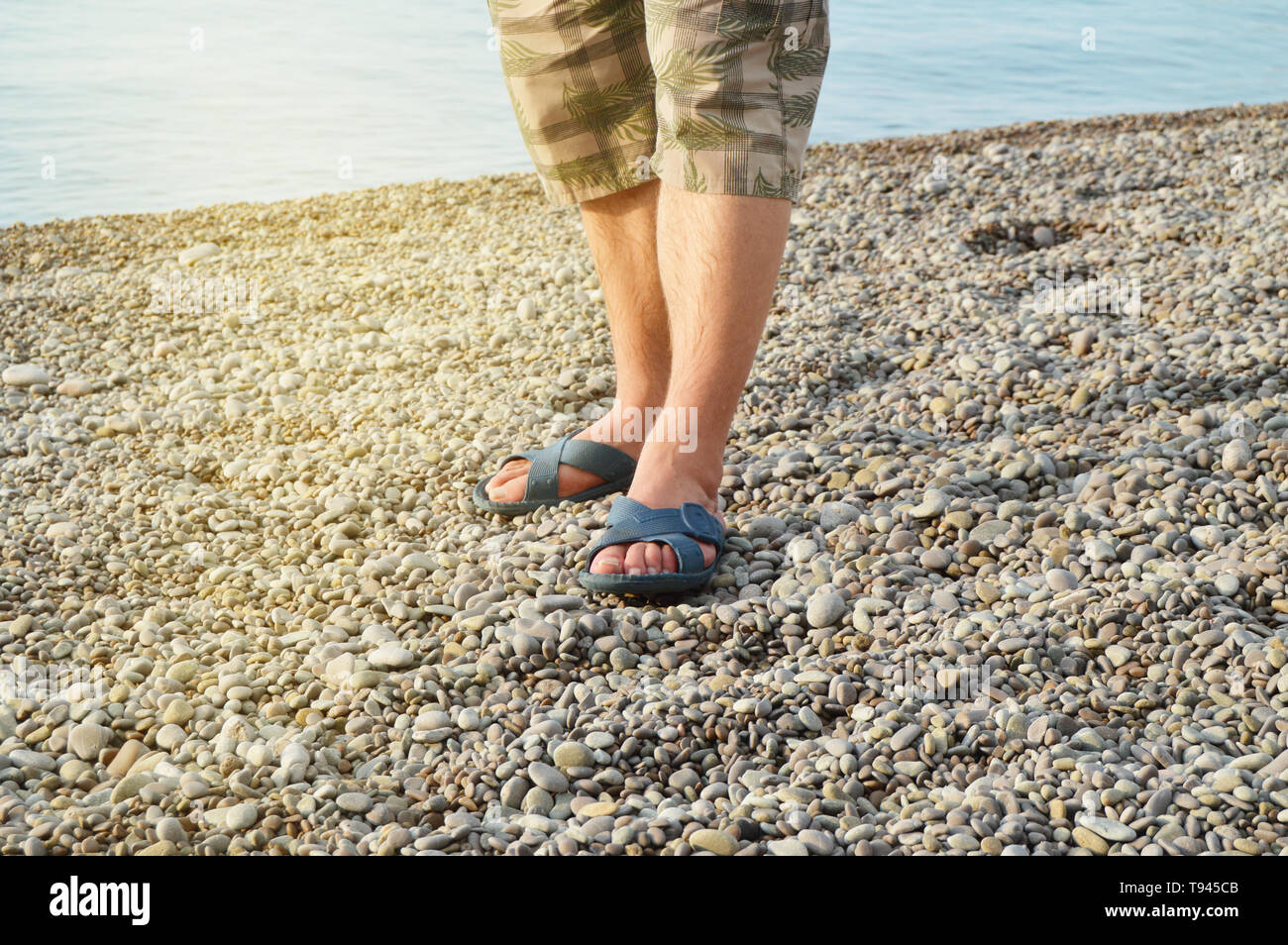 Hombre de pies en sandalias y pantalones cortos, un hombre de pie sobre la  playa, en una playa de guijarros, amanecer en la mañana Fotografía de stock  - Alamy