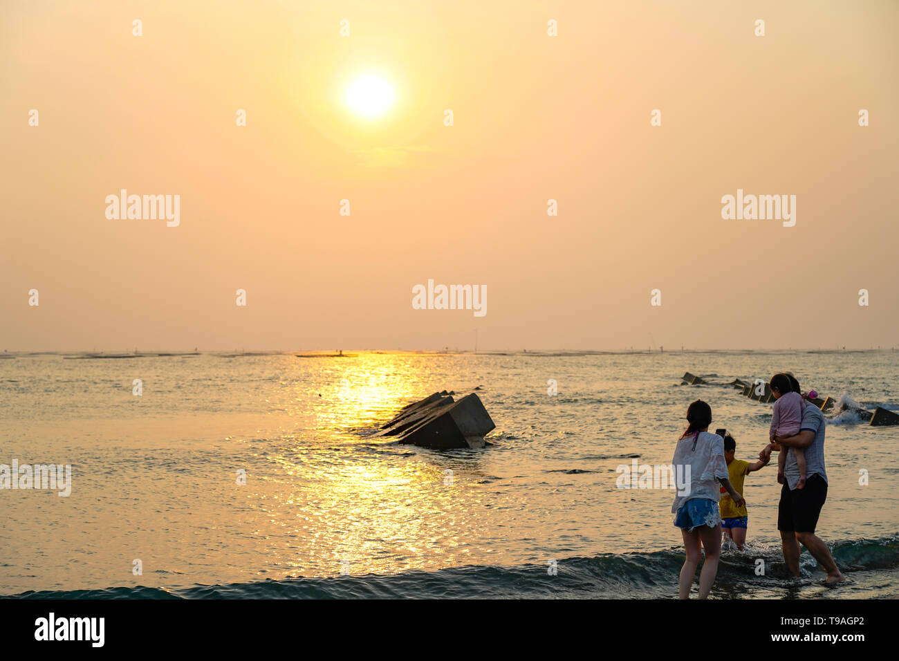 Vista de la playa con un amarillo brillante puesta de sol en el horizonte con una familia de Asia jugando en el agua. Foto de stock