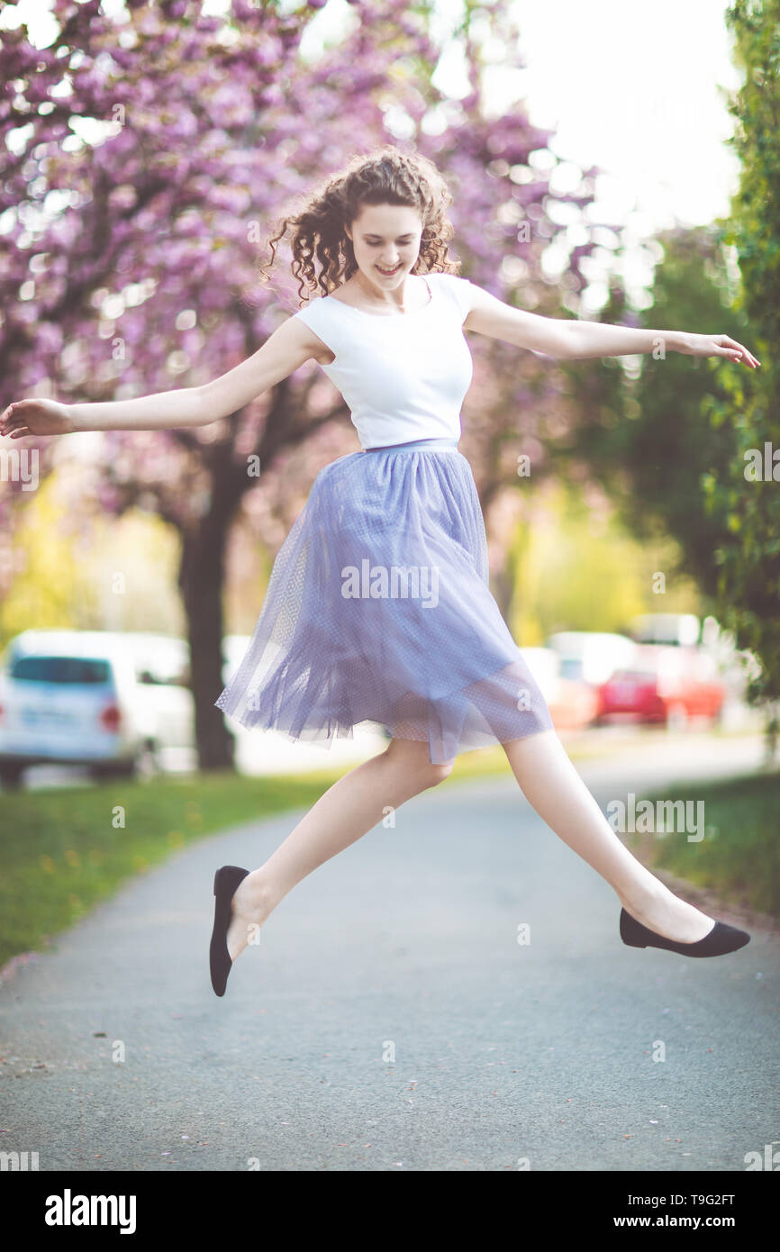 Joven con el cabello rizado en una camiseta blanca y violeta falda de baile  y saltar bajo los florecientes árboles sakura. El tiempo de primavera  Fotografía de stock - Alamy