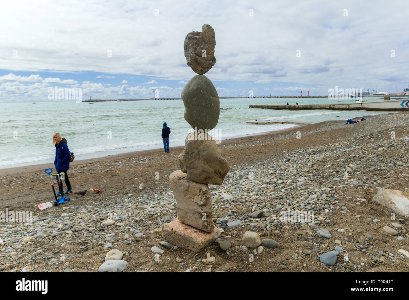 SOCHI, Rusia, 18 de abril de 2019 - pirámide de piedras en la orilla del mar Negro y la gente caminando en la playa en tiempo nublado Foto de stock