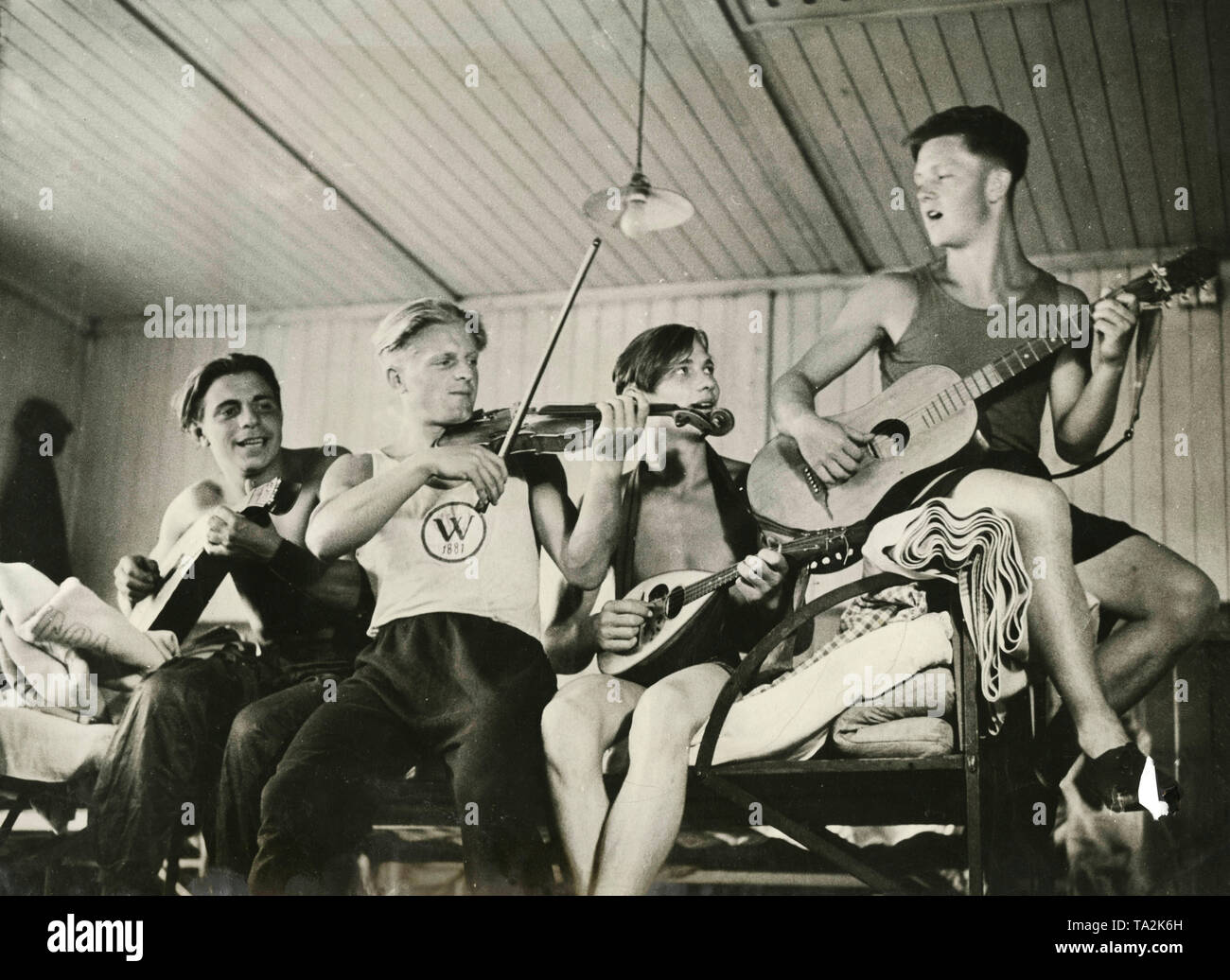 Cuatro jóvenes tocando música en una cama en un dormitorio. Están en un campamento de trabajo del Freiwilliger Arbeitsdienst en el Mar del Norte. Foto de stock