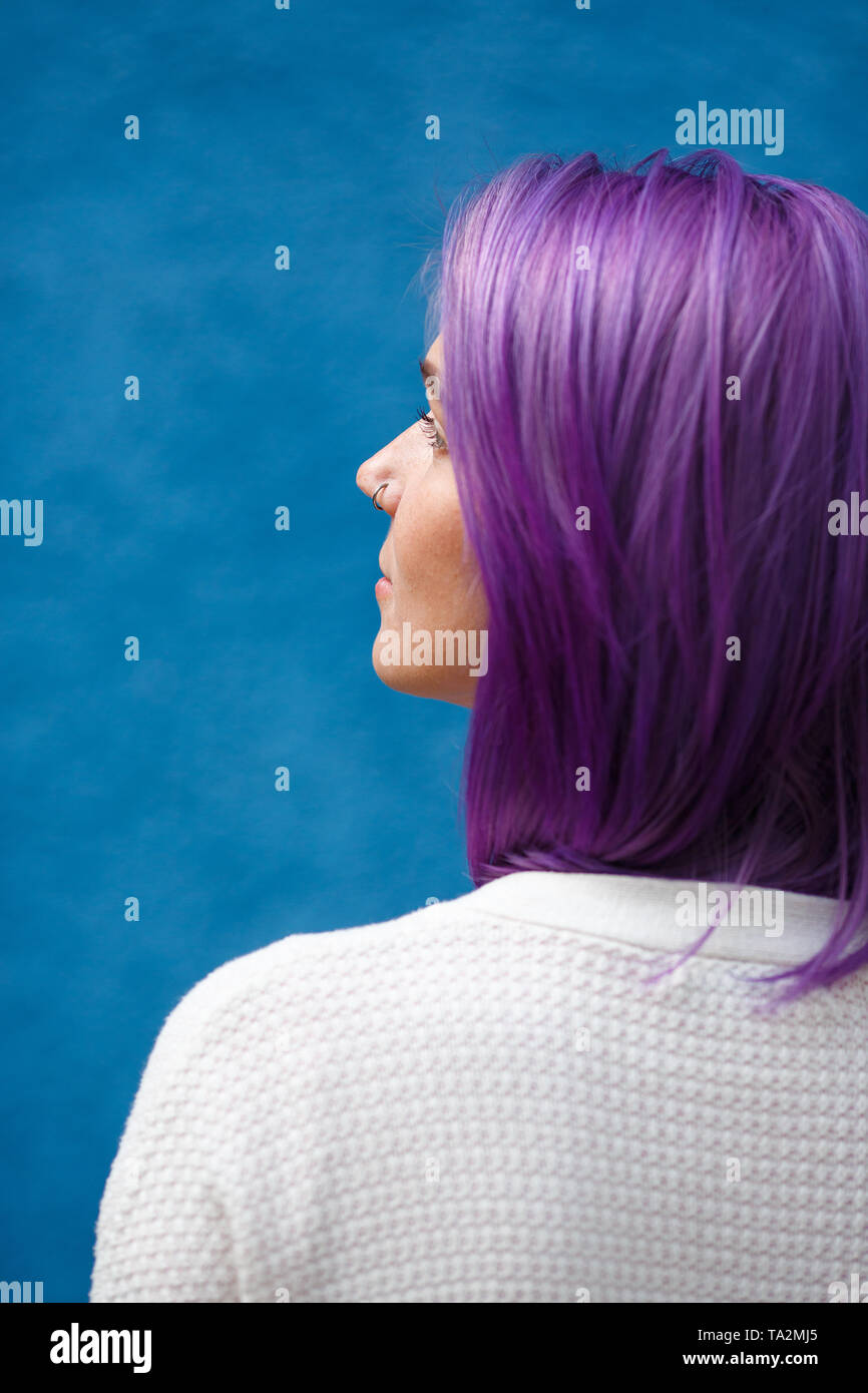 Una joven con cabello púrpura está media vuelta desde atrás sobre un fondo azul y mira hacia arriba y hacia el lado, blanco chaqueta tejida Foto de stock