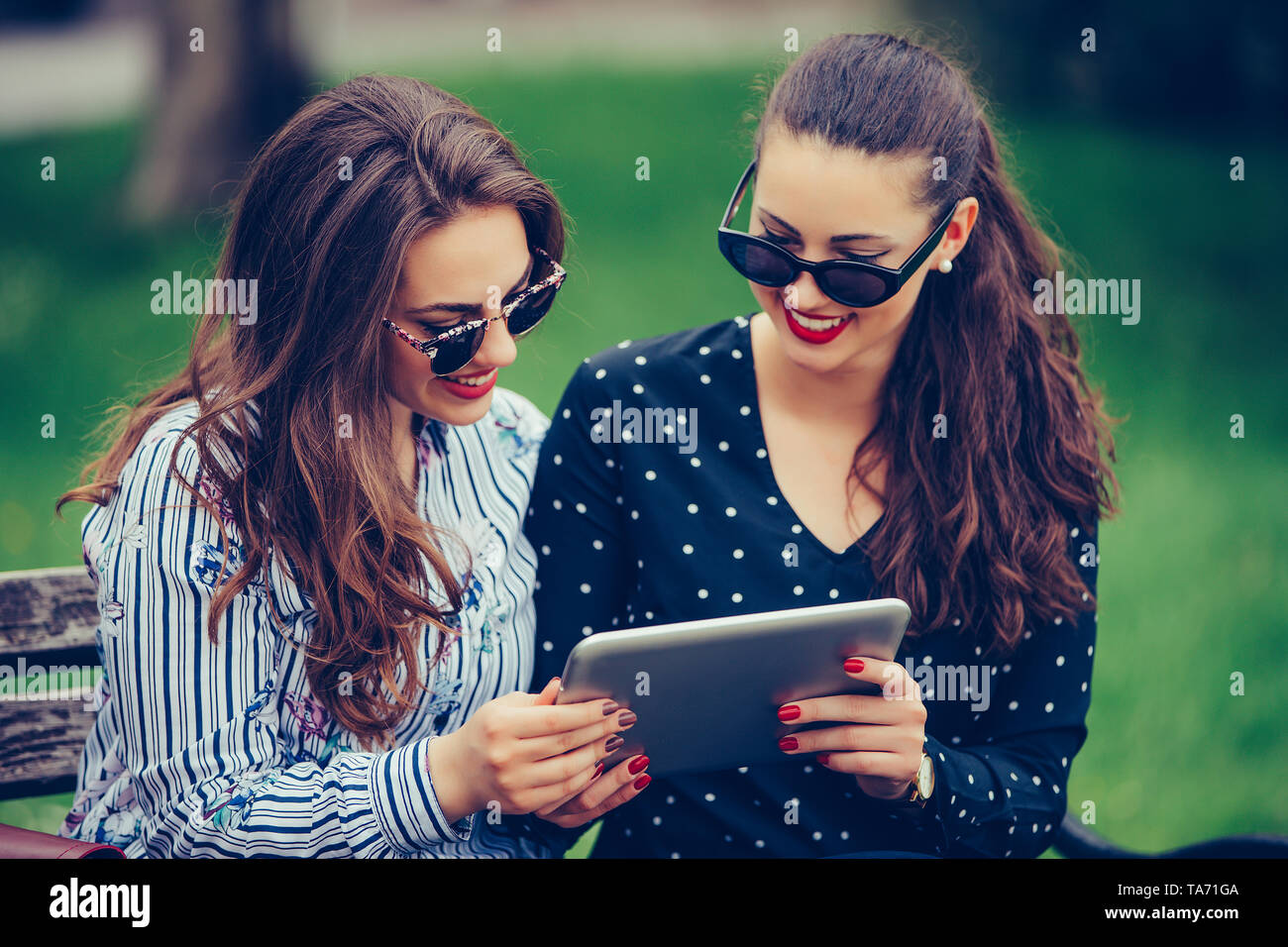 Dos mujeres hermosas riendo viendo contenido multimedia juntos en una tableta digital, sentados en un banco en el parque. - Imagen Foto de stock