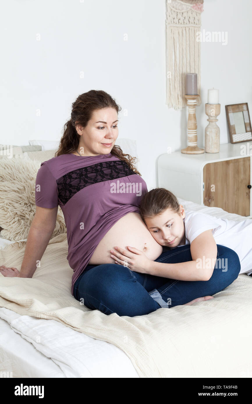 Madre embarazada con la barriga Desnuda sentada en la cama con su hija adolescente que escucha el bebé, dormitorio Fotografía de stock foto