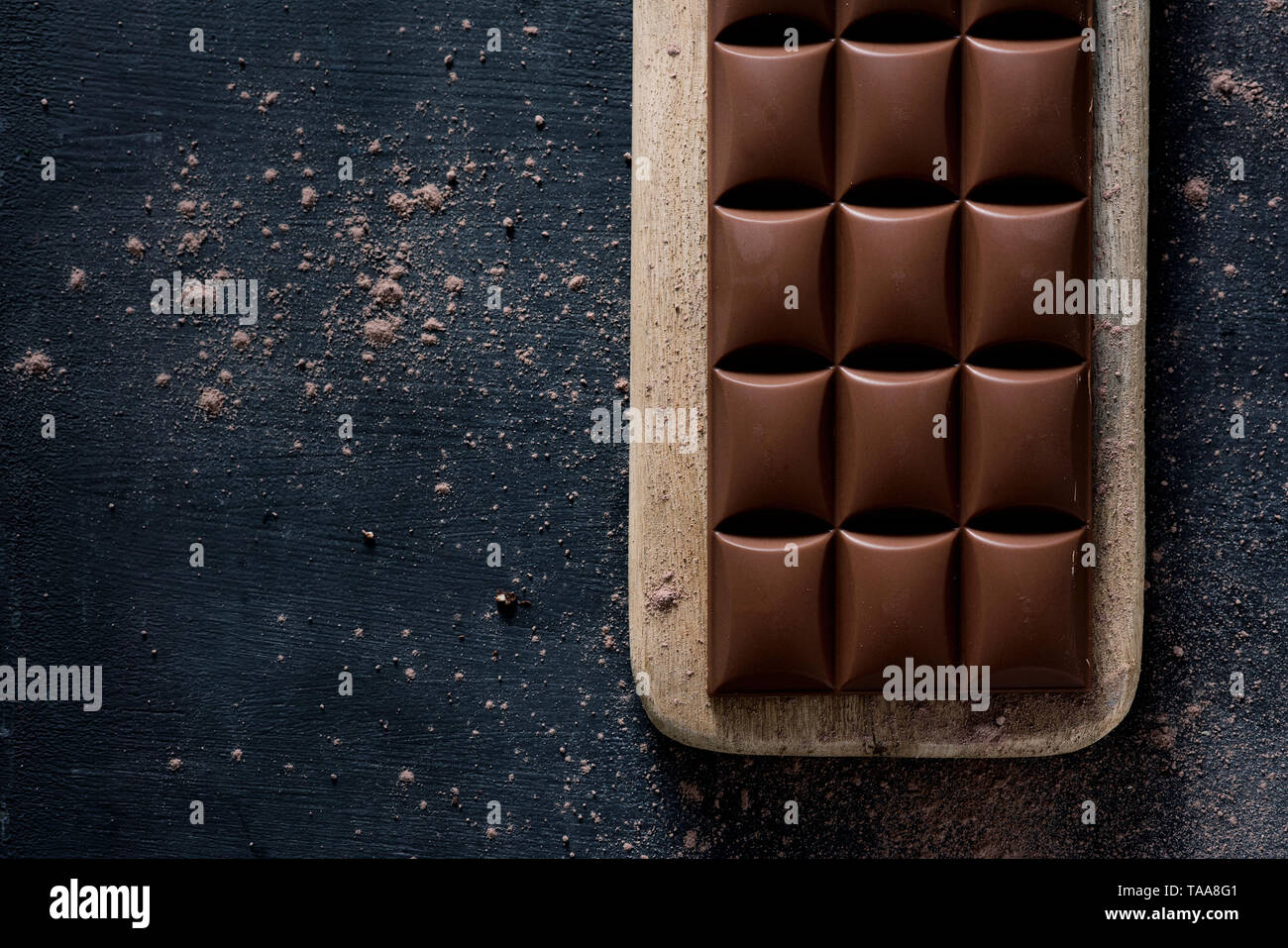 Un alto ángulo de visualización de una barra de chocolate en una placa de madera, colocado sobre una mesa de madera rústica oscura espolvoreada con cacao en polvo, y algún espacio en blanco de la le Foto de stock