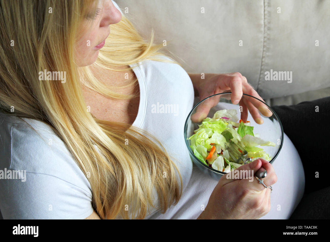 Un saludable joven embarazada con largo pelo rubio es sentarse y descansar mientras come una ensalada verde frondosa. Foto de stock