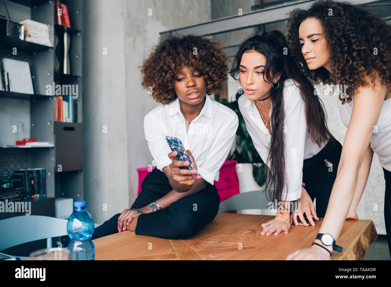 Tres mujeres jóvenes multirracial mirando el smartphone en la moderna oficina coworking - compañerismo, interacción, reserva Foto de stock