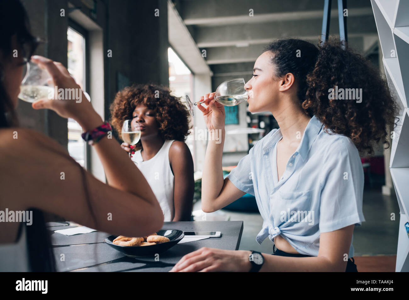 Jóvenes mujeres multirraciales sentado en un moderno restaurante bebiendo juntos vino - amistad, alegre, comunicación Foto de stock