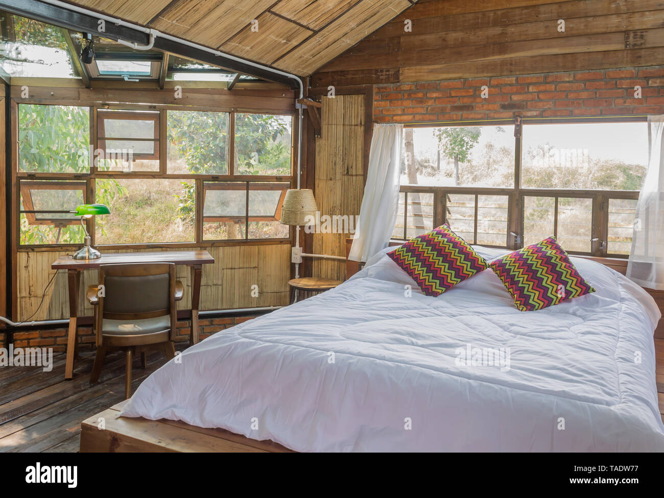 La cama blanca con almohadas y mesa de trabajo en la Sala de diseño de  interiores del país. Habitación de diseño interior incluyen silla y lámpara  y ventanas y cortinas. Interior de