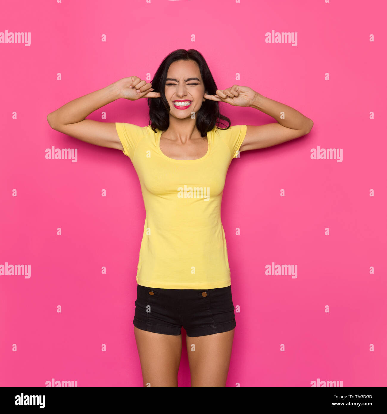 Hermosa joven en camisa amarilla y pantalón negro es la celebración de  dedos índices en los oídos, cerrar los ojos y sonriendo. Tres cuartos de  longitud studio shot Fotografía de stock -