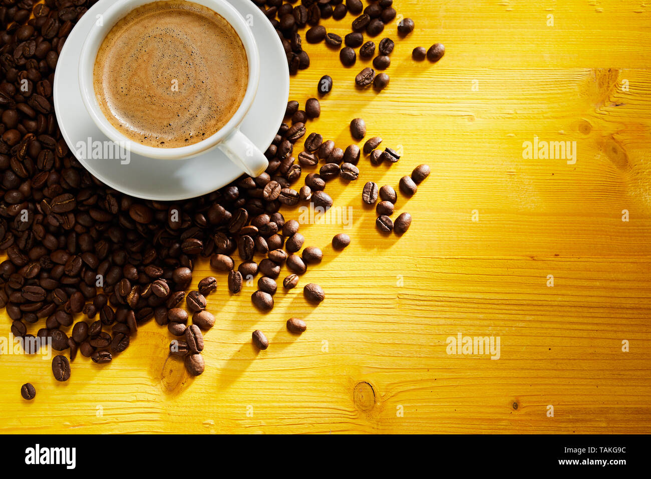 Una deliciosa taza de café capuchino espumoso de café tostado en grano dispersos en la esquina en coloridos de madera amarilla con espacio de copia Foto de stock