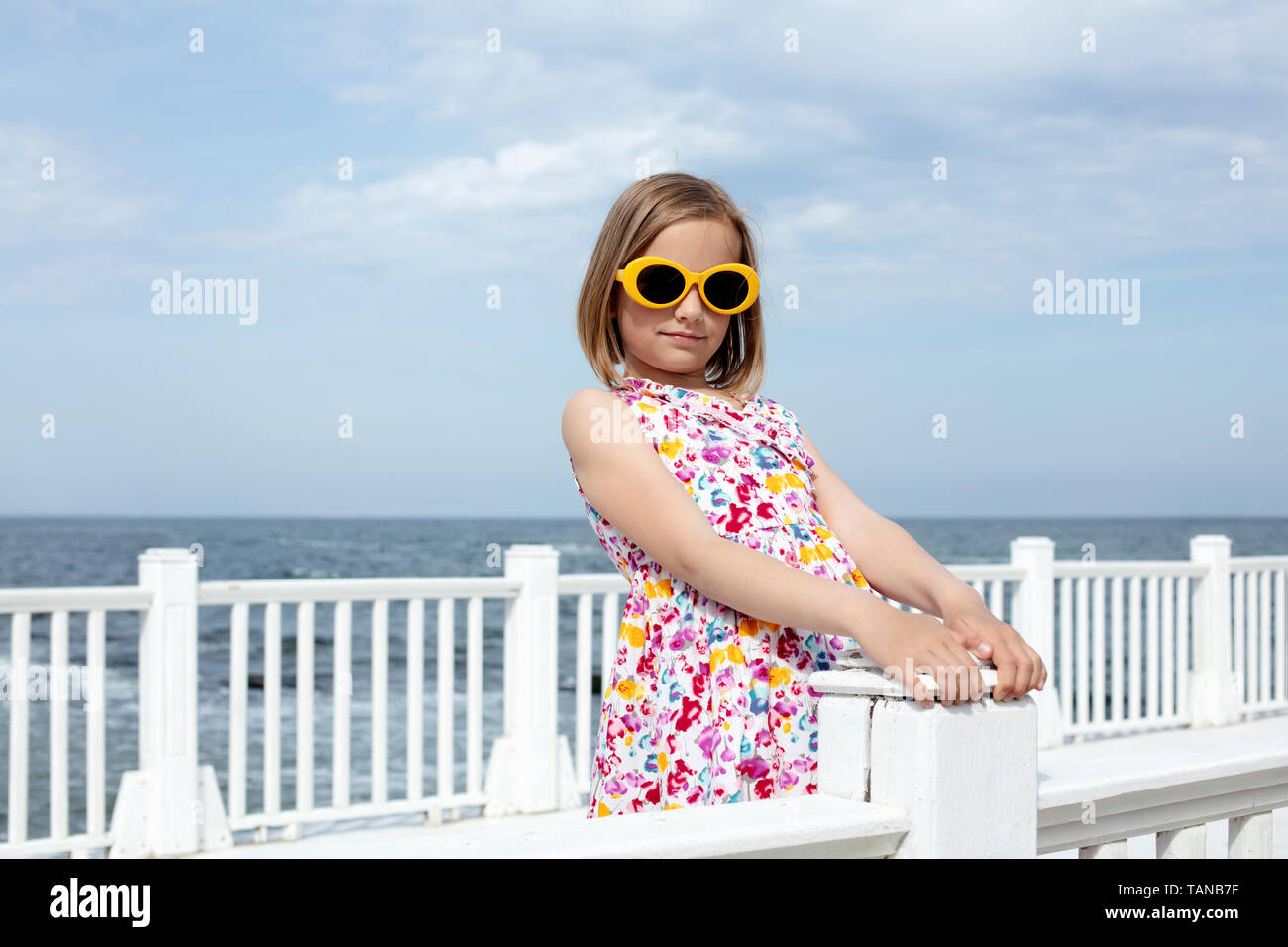 Pequeño (8 años) bastante alegre niña en un vestido con estampados florales  en amarillo y gafas de sol se encuentra en un muelle blanco de madera. El  enfoque selectivo Fotografía de stock -