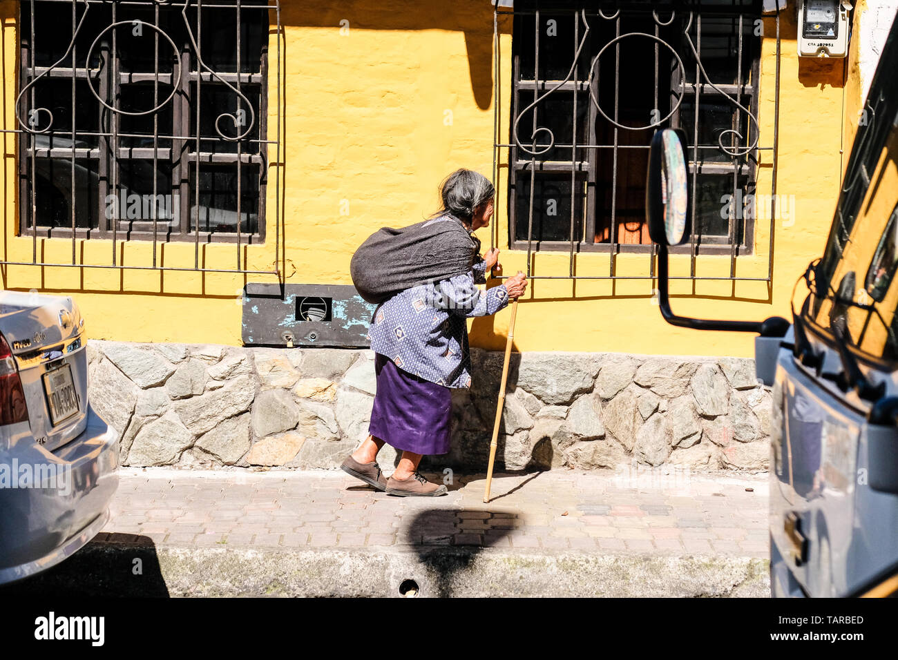 Una vieja mujer indígena ecuatoriano con bolsa pesada sobre su espalda camina despacio en la acera Foto de stock
