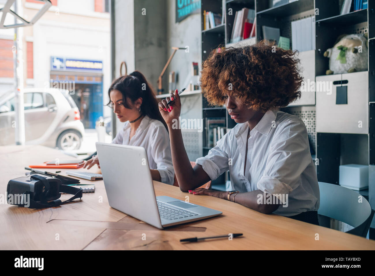 Dos jóvenes mujeres multirracial de escribir y trabajar con dispositivos digitales modernos en espacio de coworking - freelance, interacción, concentración Foto de stock
