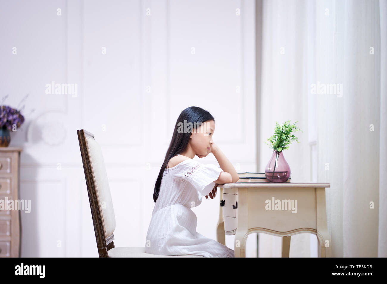 Triste poco de Asia chica con largo cabello negro sentado en una mesa en su habitación pensando con la mano en la barbilla Foto de stock