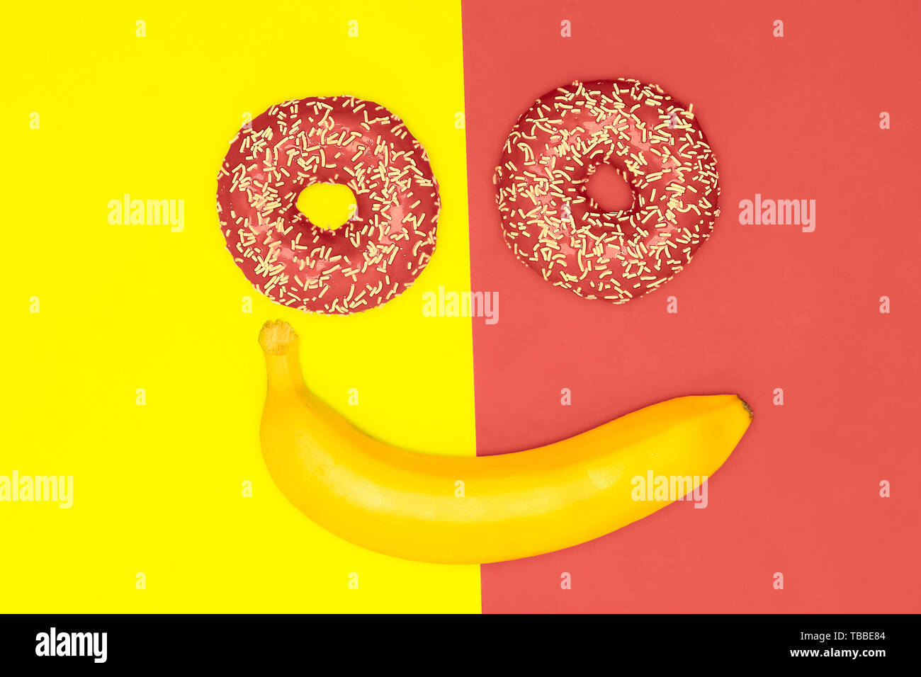 Buen humor, sonrisas concepto de comida. Las rosquillas y banana en la forma de una cara sonriente. Coral vivo laicos plana Foto de stock