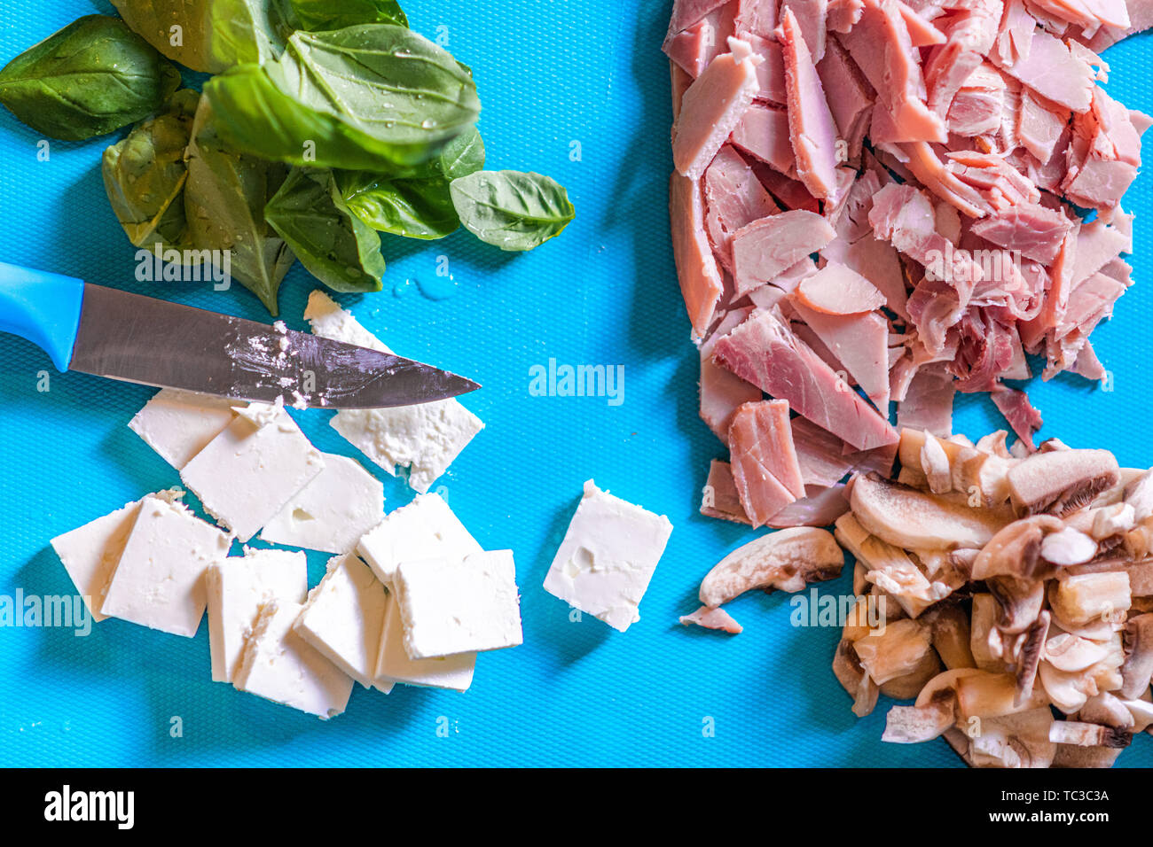 Cortar ingredientes como jamón, setas, quesos y albahaca sobre una tabla de cortar de plástico azul con un cuchillo afilado corta Foto de stock