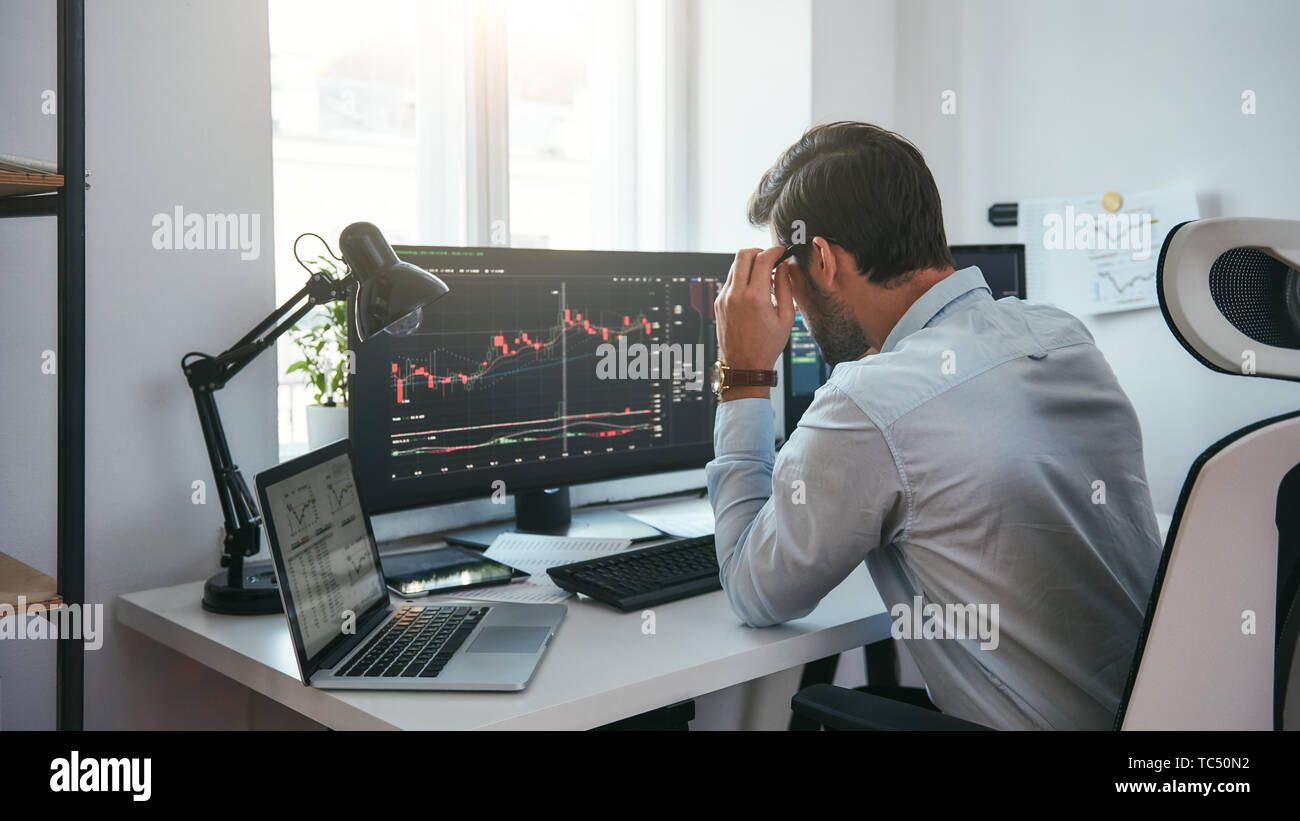 Proceso de trabajo. Smart Trader macho mirando gráficos en varias pantallas  de ordenador y analizar los datos mientras está sentado en su oficina  moderna. Bolsa de Valores. Concepto de comercio. Concepto de