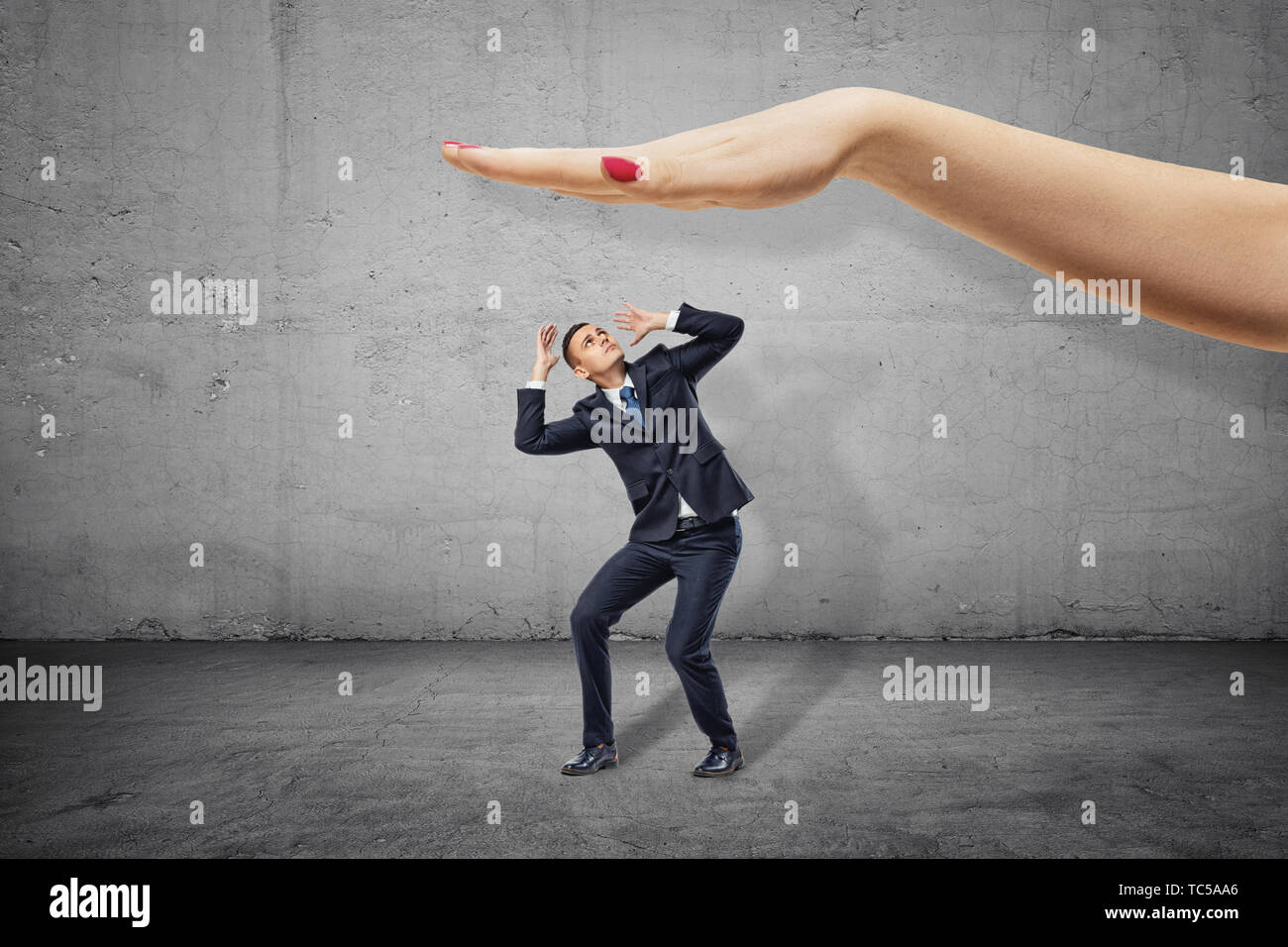 Poco empresario cubriendo la cabeza con las manos y mirando por encima de la mano del gran mujer dispuesta a swat él. Foto de stock