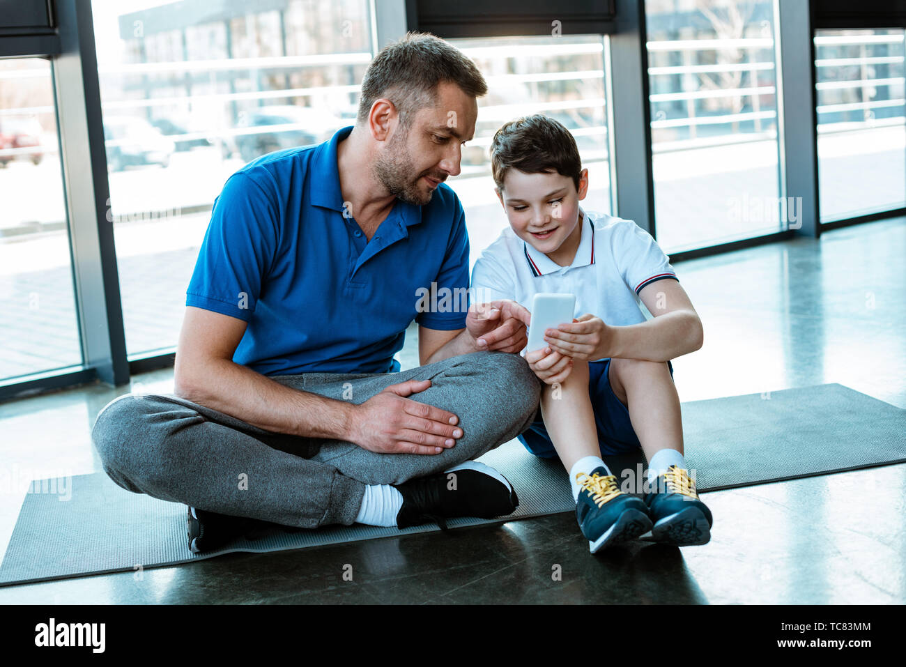 Padre e hijo sentado en colchoneta de fitness en el gimnasio y utilizando el smartphone Foto de stock