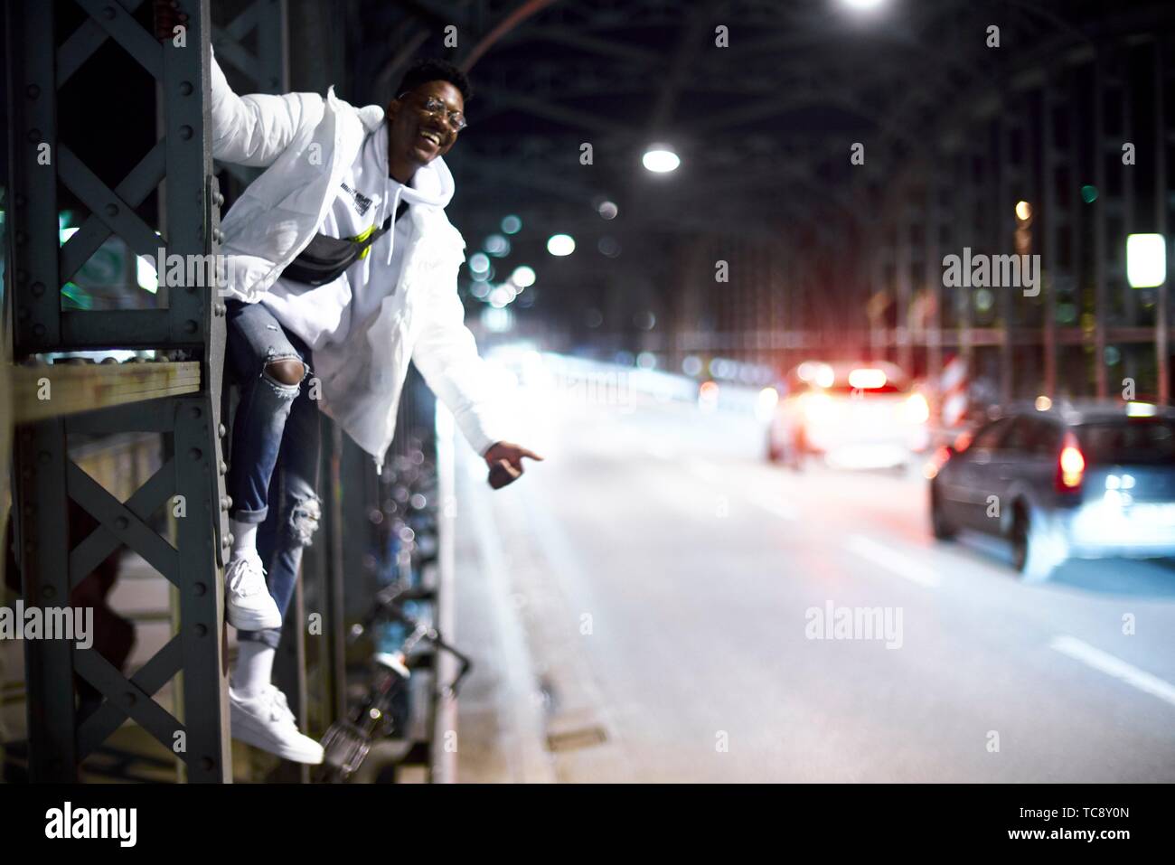 Loco joven gracioso hombre tratando de autostop, colgando sobre metal construcción de puente, Hacker Haccurbrücke bridge, en la noche en la ciudad, al lado de la calle, en Foto de stock