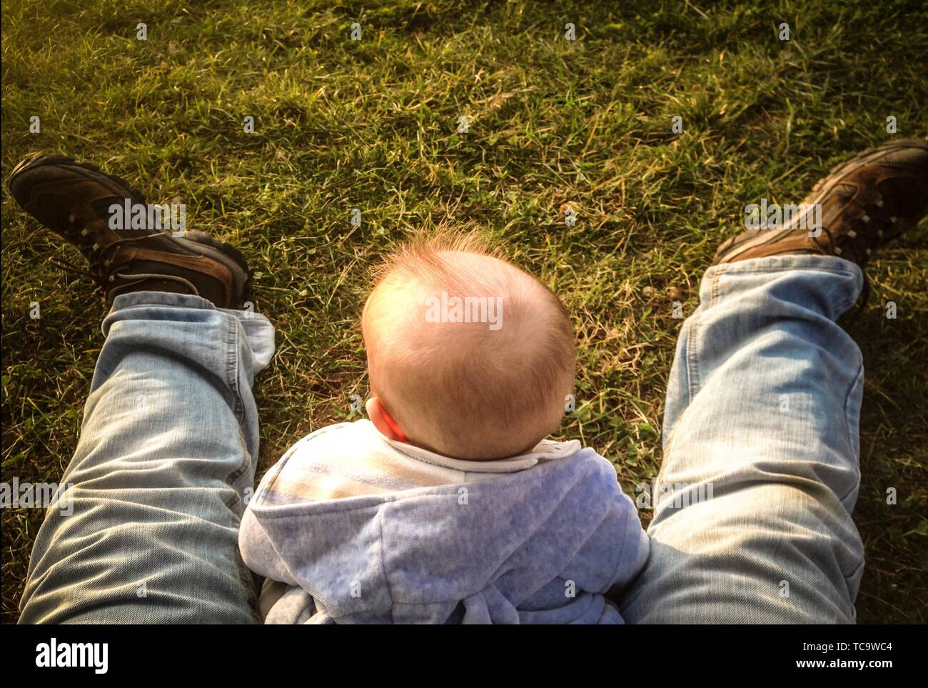 Baby Boy sentado sobre hierba entre sus padres las piernas. Los padres teniendo cuidado de bebés concepto. Foto de stock