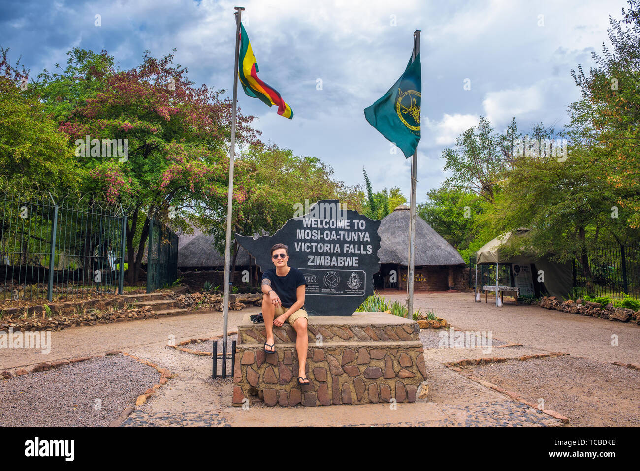 Turista se sienta en el cartel de bienvenida colocado en la entrada de Victoria Falls, Zimbabwe Foto de stock