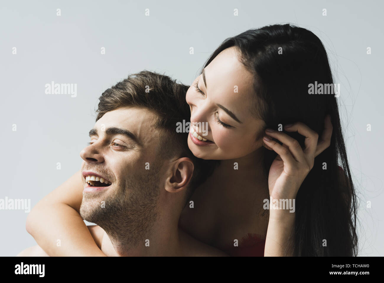 Feliz mujer asiática en rojo bra abrazan sonrientes guapo novio sobre gris Fotografía de stock
