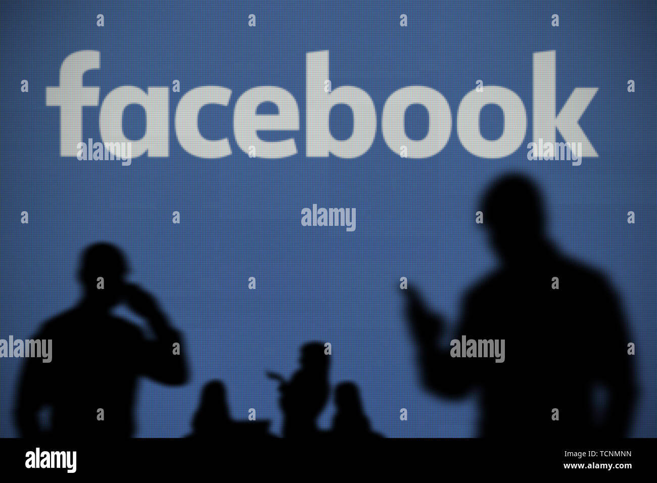 El logotipo de Facebook es visto en una pantalla LED en el fondo mientras una silueta persona utiliza un smartphone en primer plano (uso Editorial solamente) Foto de stock