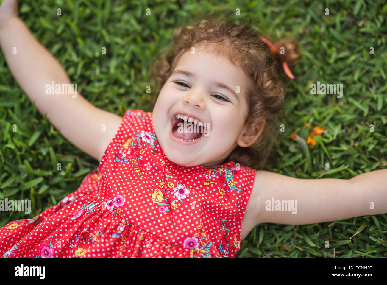 Feliz poco sonriente Niño Niña sentar en la hierba de parque con vestido rojo. Foto de stock