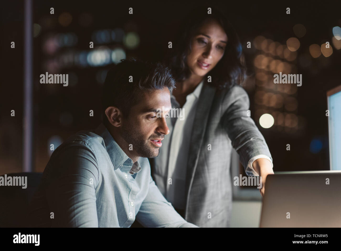 Dos jóvenes empresarios mediante un portátil juntos mientras trabajan horas extraordinarias en un escritorio de oficina en la noche Foto de stock