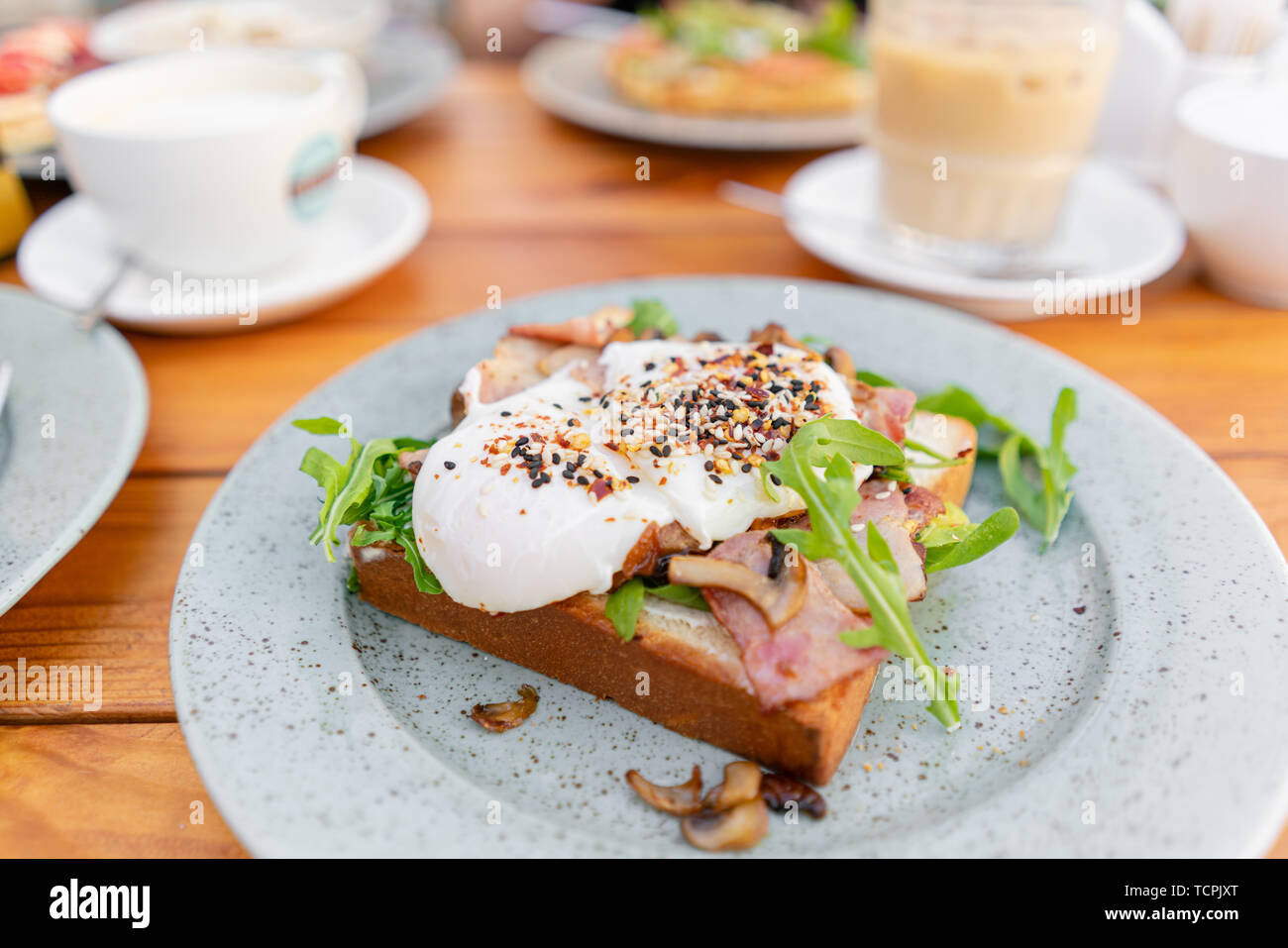 El desayuno y el café en la cafetería de verano. Sandwich en un panecillo brioche con tocino, champiñones, rúcula y huevo escalfado Foto de stock