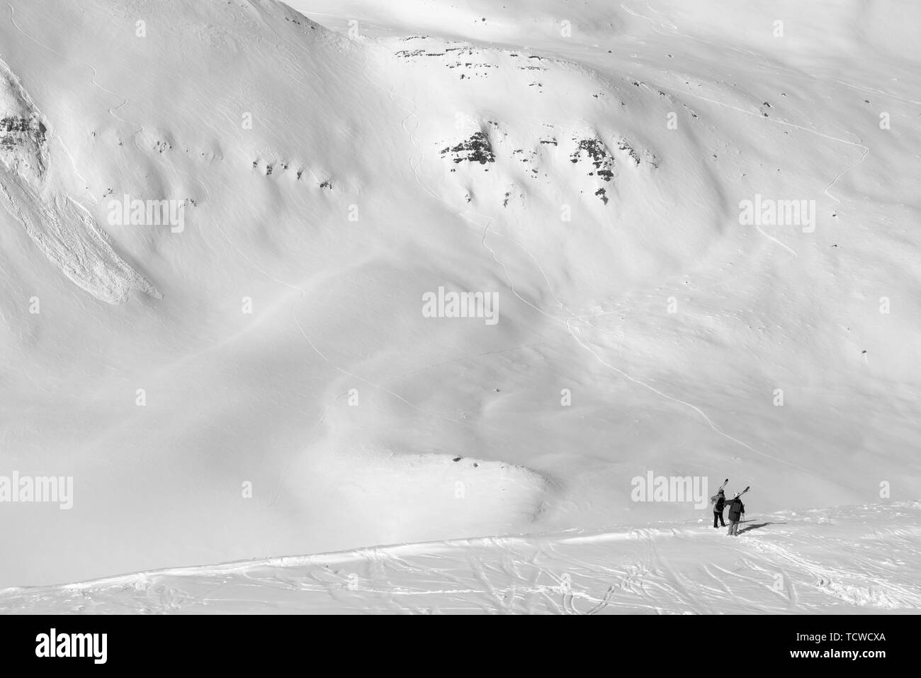 Dos esquiadores con esquíes sobre su hombro y fuera de pista pendiente con huellas de esquíes, tablas de snowboard y avalanchas. Montañas del Cáucaso en sol de invierno frío día, Ge Foto de stock