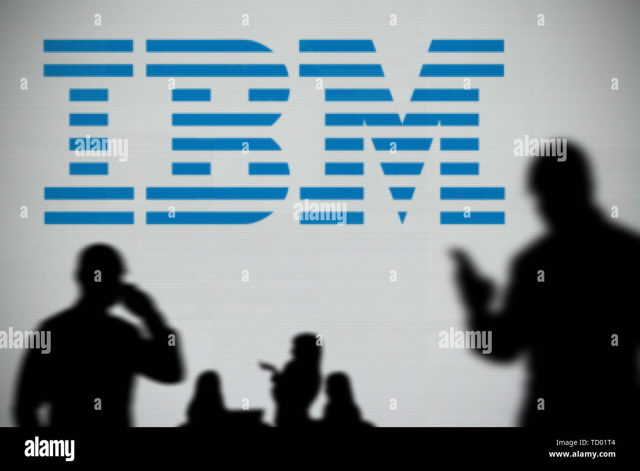 El logotipo de IBM es visto en una pantalla LED en el fondo mientras una silueta persona utiliza un smartphone en primer plano (uso Editorial solamente) Foto de stock