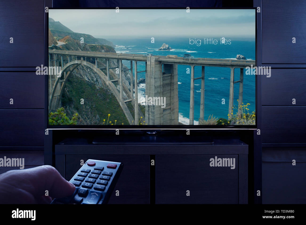 Un hombre apunta un control remoto de la televisión en la televisión que muestra la gran pantalla del título principal LITTLE LIES (uso Editorial solamente). Foto de stock