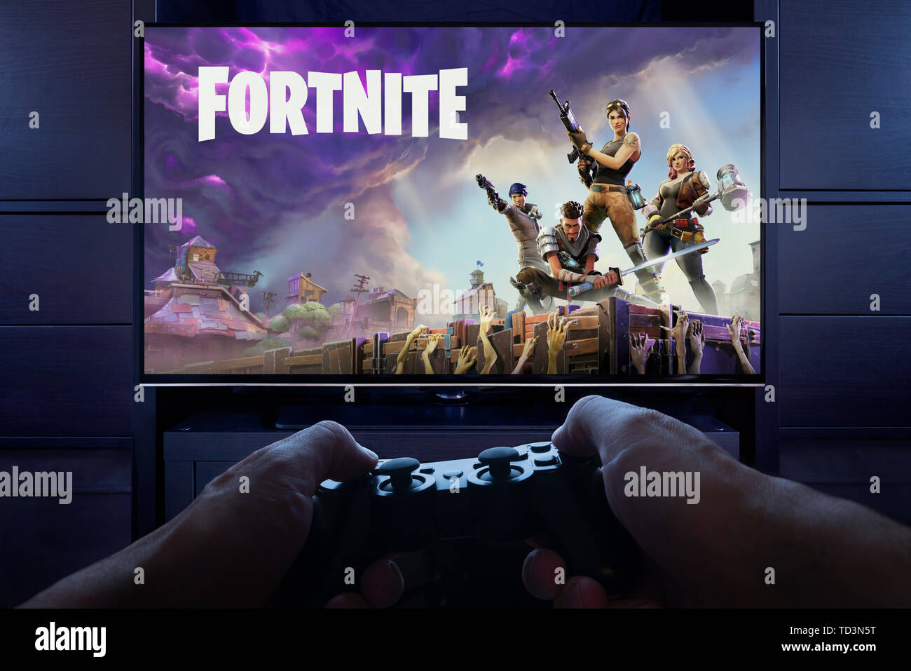 Con controlador de Playstation en la mano, un hombre se prepara para reproducir Fortnite: salvar el mundo como preludio de la pantalla de carga en visto en un televisor (sólo para uso editorial). Foto de stock