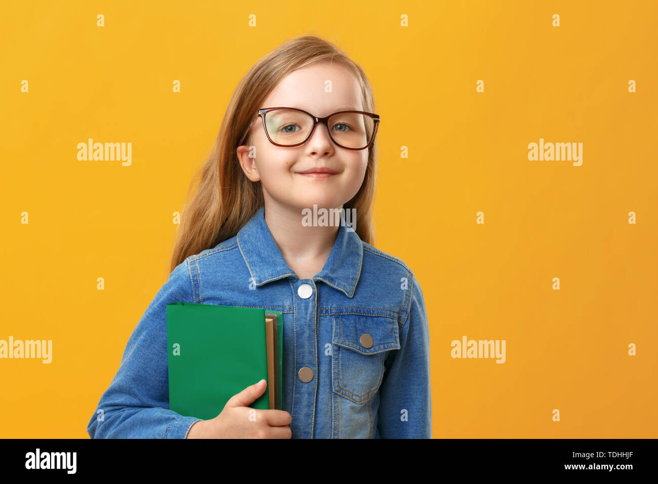 Poco estudiante chica en denim shirt sobre un fondo amarillo. Un niño con gafas, sostiene un libro. El concepto de la educación y de la escuela. Foto de stock