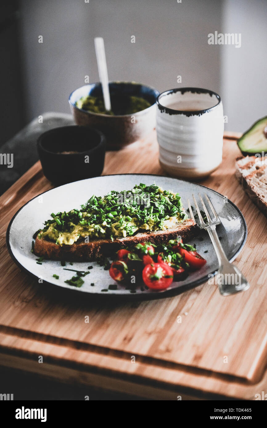 Vegan saludable desayuno en verano. Aguacate tostadas de pan de masa fermentada con cebollino, cilantro y tomates cereza con taza de café sobre la plancha de madera. Vegeta Foto de stock