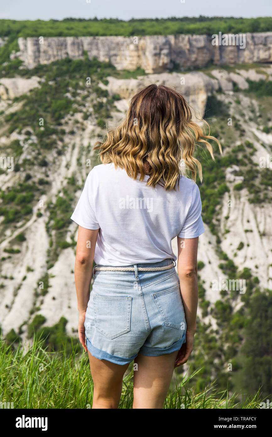 Una chica rubia con el cabello ondulado en una camiseta blanca está con su  espalda y mira a las montañas. La libertad y el concepto de viaje  Fotografía de stock - Alamy