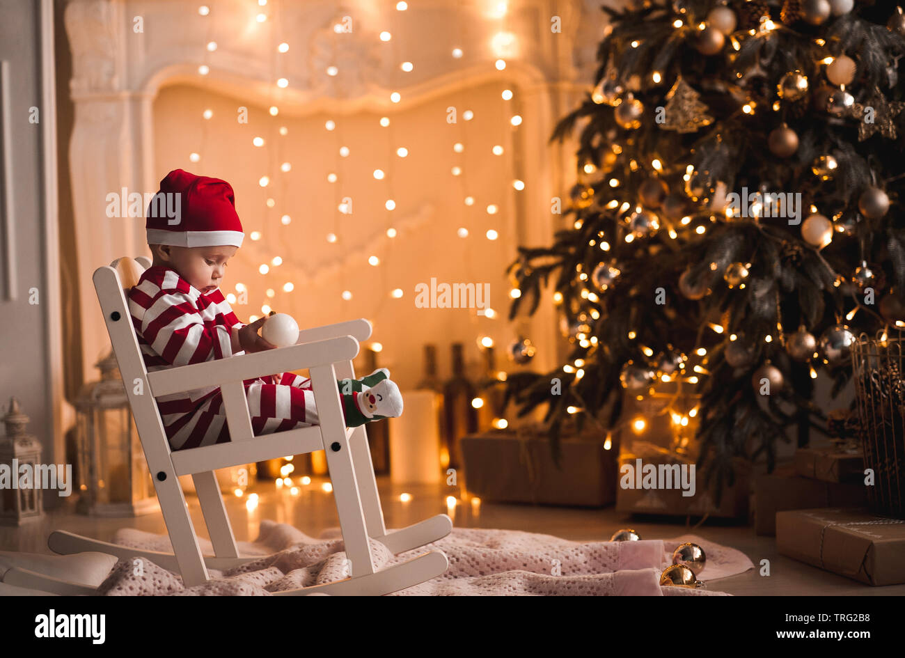 Bebé de 1 año de edad vistiendo traje de santa claus sentado en una  mecedora con árbol de Navidad y luces de fondo en la habitación. Feliz  Navidad. Holiday seaso Fotografía de