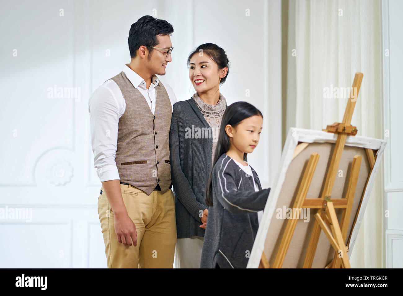 Bonita chica asiática con largo cabello negro haciendo una pintura sobre lienzo mientras los padres de pie detrás mirando, se centran en los padres. Foto de stock