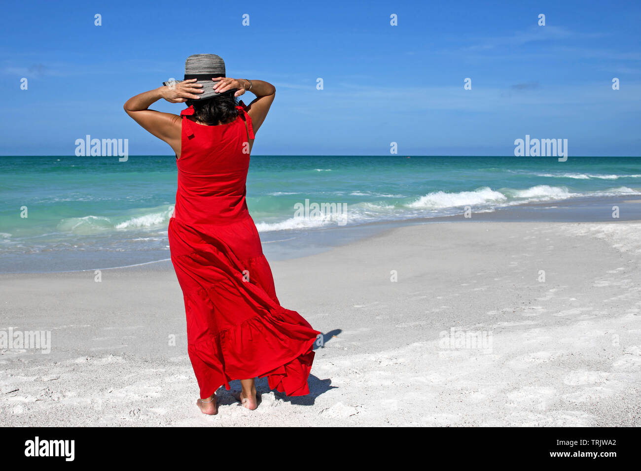 Bella en vestido rojo de pie en la playa, sosteniendo su sombrero en la cabeza para mantenerlo de soplar lejos Fotografía de stock - Alamy