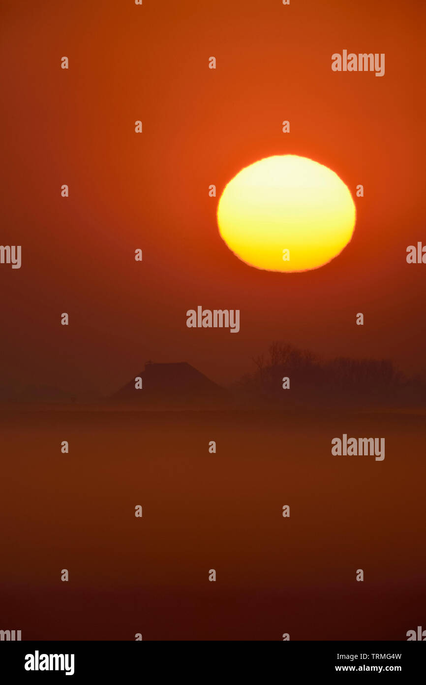Impresionante Amanecer sobre un caserío y algunos campos cubiertos de neblina temprano por la mañana, el cielo de color rojo, sun loooks como una enorme bola de fuego, Europa. Foto de stock