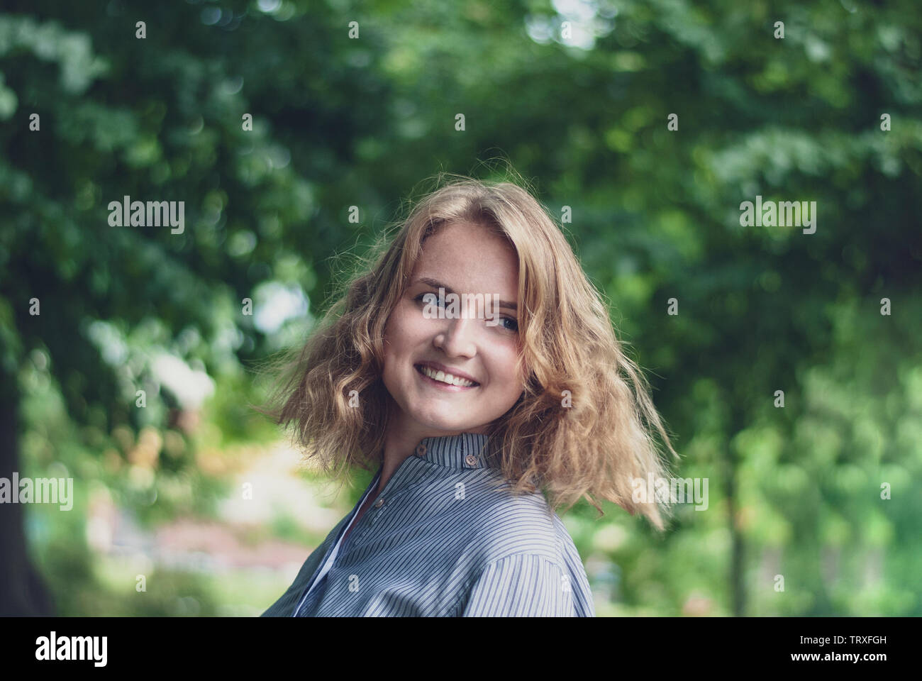 Feliz atractiva mujer rubia mirando hacia atrás sobre su hombro con una cálida sonrisa feliz al aire libre en un parque enselvado Foto de stock