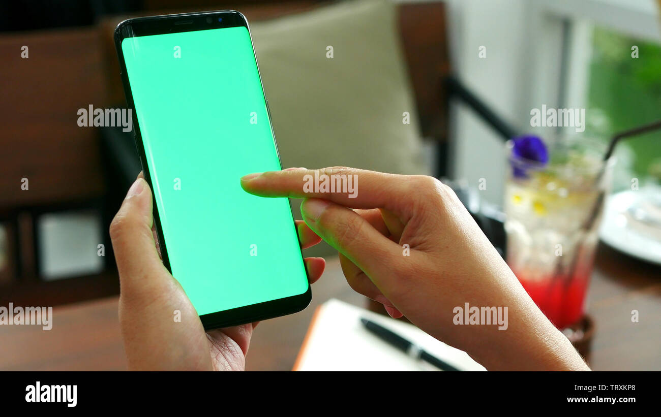 Mujer mano sujetando el teléfono inteligente móvil con pantalla verde en blanco, utilice la punta del dedo la diapositiva en pantalla verde en la cafetería con bebidas a fondo Foto de stock