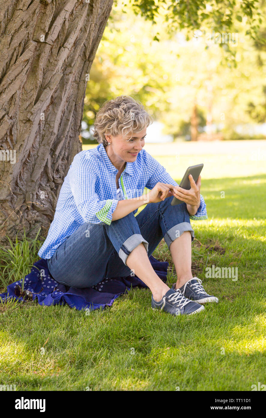 Feliz y sonriente, de mediana edad mujer madura, utilizando un equipo de tableta digital e-reader al aire libre en un parque. Ella está tocando la pantalla. Foto de stock